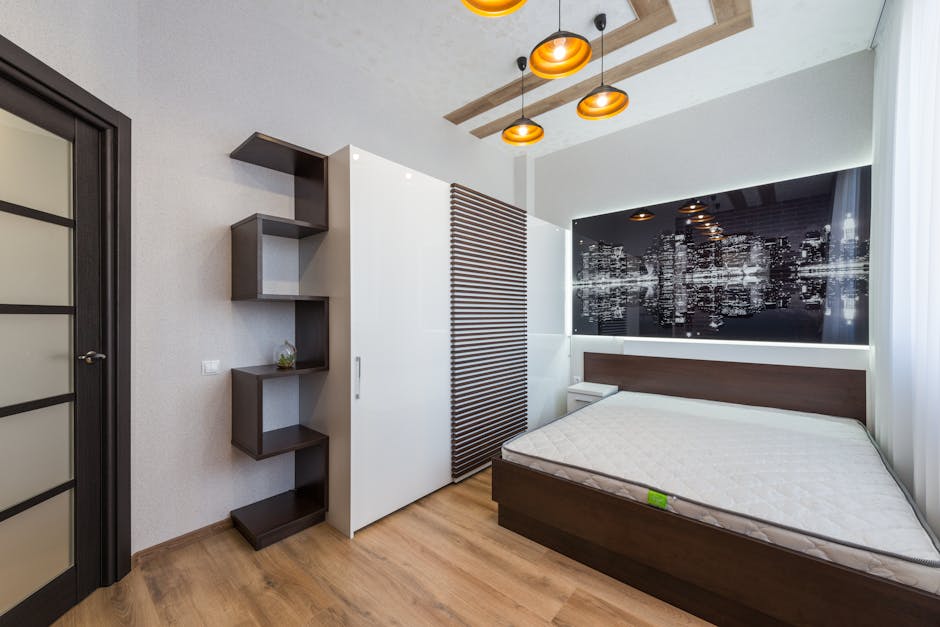 Bed with white mattress placed at wall with decorative picture near white wardrobe and black shelves in bedroom with door