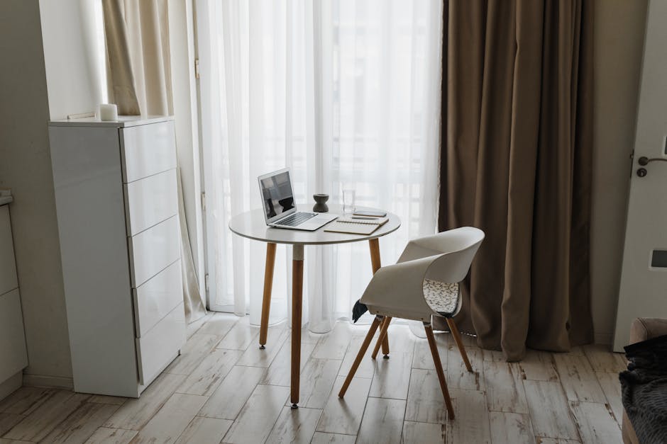 Minimalist home office setup with a laptop, table, and chair by a window for a serene workspace.