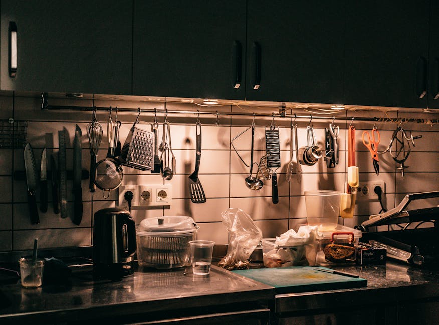 Kitchen counter with cupboards and assorted utensils hanging above table top with dishware teapot and cutting board in dark kitchen