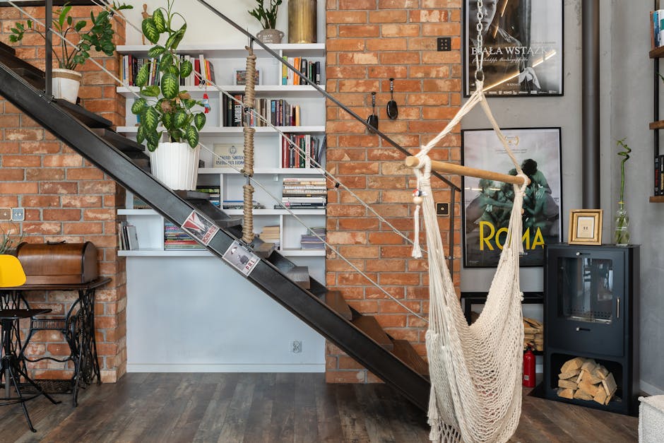 Cozy industrial loft interior featuring a hammock, modern staircase, brick wall, and bookshelf.