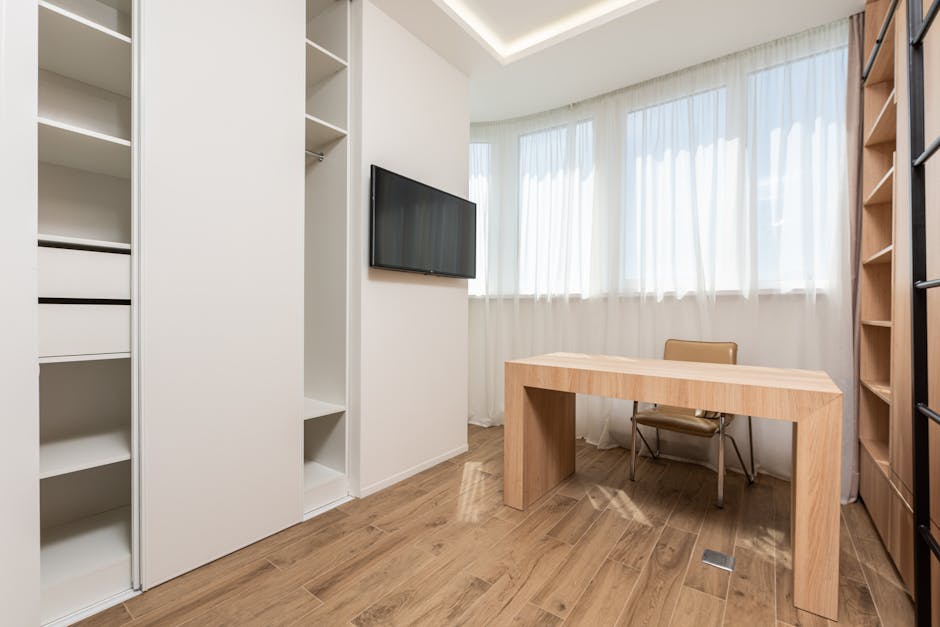 Chair at wooden table near wall with window and TV between opened empty wardrobes in spacious room with minimalist interior