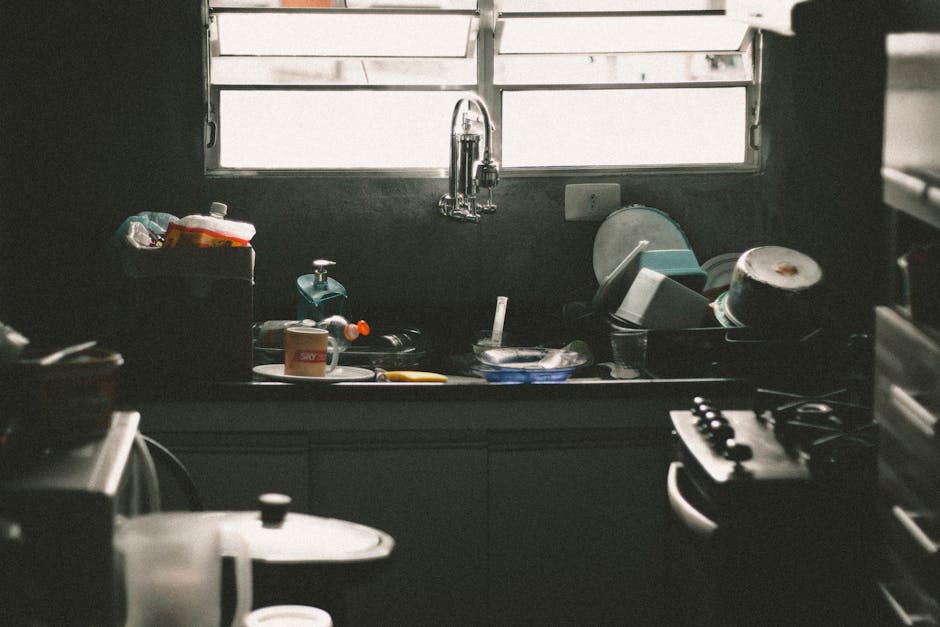 A cluttered kitchen sink filled with dirty dishes and utensils near a window, creating a messy home interior scene.
