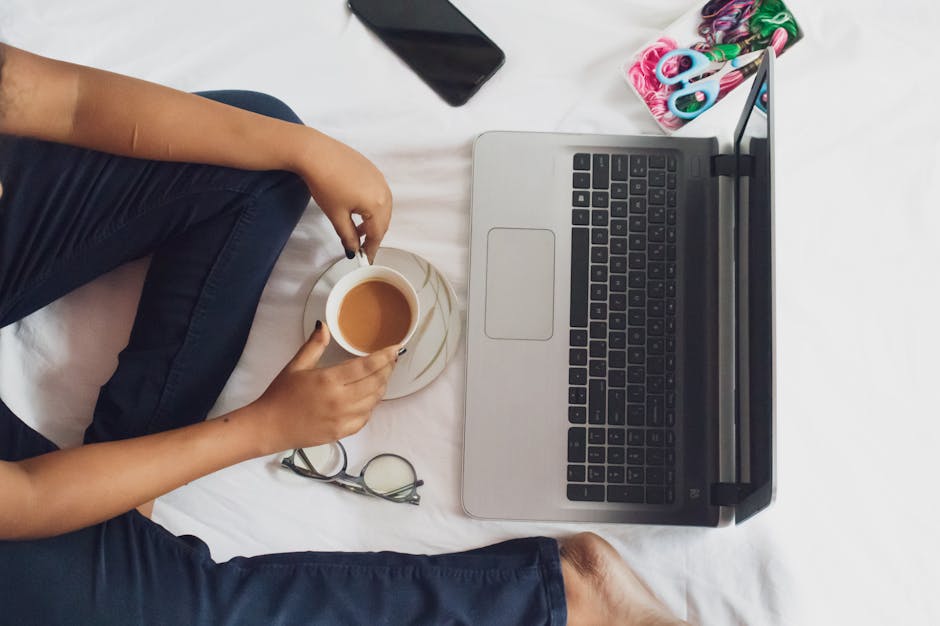Comfortable remote work setup featuring a laptop, coffee, and essential tools on a white bed.
