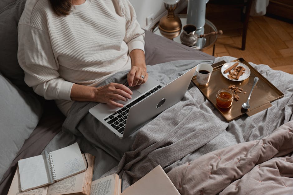 Person working on a laptop in bed with breakfast tray, books, and a warm ambiance.