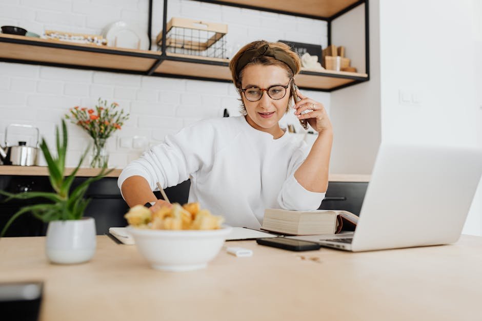 Adult woman in a white sweatshirt working from home office with laptop and phone, in a stylish kitchen.